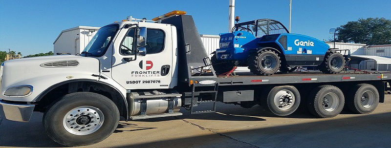 A parked flatbed tow truck towing a Genie® telehandler near a white building on a clear sunny day.